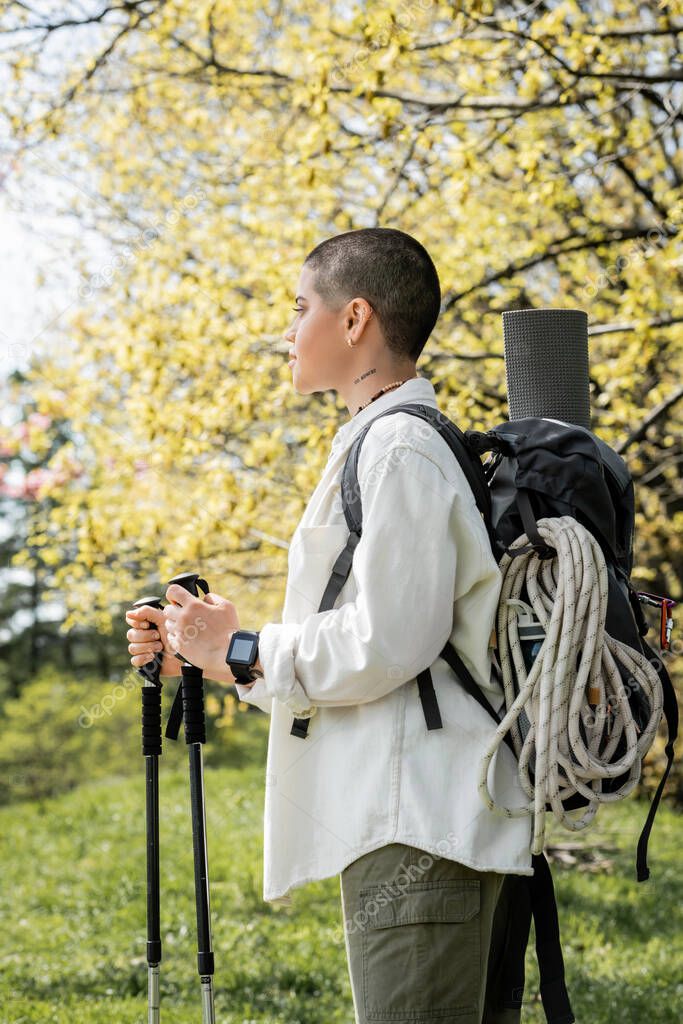 Side view of young short haired and tattooed female hiker with backpack and climbing rope holding trekking poles and walking on blurred nature, independent traveler embarking on solo journey