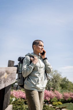 Smiling young short haired female tourist with backpack and map talking on smartphone and looking away while standing with blurred nature and blue sky at background, Translation of tattoo: love clipart