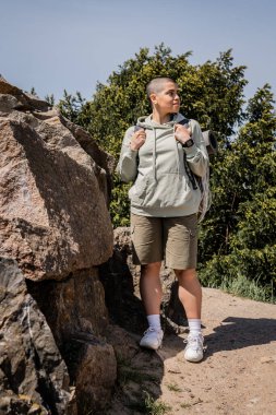 Young short haired female tourist with fitness tracker and backpack looking away while standing near stones with nature and blue sky at background, exploring new horizons, Translation of tattoo: love clipart