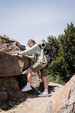 Sırt çantalı, seyahat ekipmanlı taşlara dokunan ve arka planda manzarası olan yeni ufuklar keşfeden genç, kısa saçlı turistin yan görüntüsü.