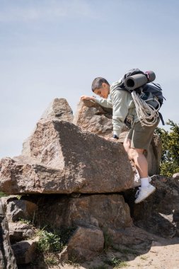 Side view of young short haired female tourist with backpack and travel equipment climbing on stones with blue sky and nature at background, exploring new horizons, summer clipart