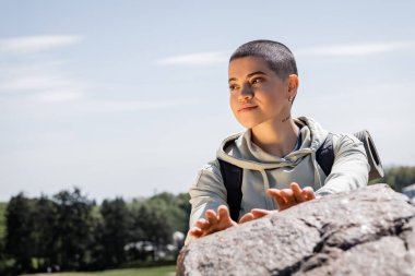 Portrait of smiling short haired female hiker with backpack looking away while standing near blurred stone with sky and landscape at background, vibrant travel experiences, summer clipart