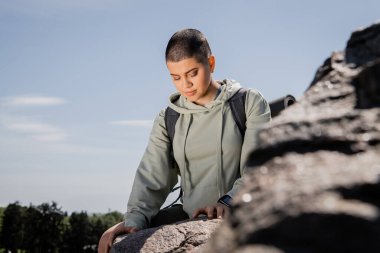Young short haired female tourist with backpack and fitness tracker standing near blurred stones with blue sky and landscape at background, vibrant travel experiences, summer clipart