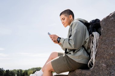 Young short haired female traveler with backpack and travel equipment using smartphone while sitting on stone with nature and blue sky at background, vibrant travel experiences, summer clipart