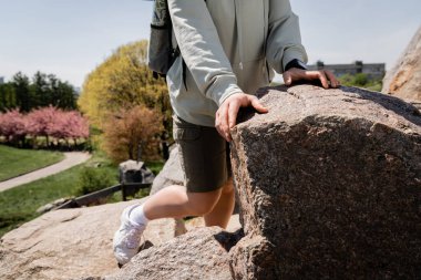 Cropped view of young female hiker with backpack and fitness tracker standing near stones on hill with blurred nature at background, vibrant travel experiences, summer clipart