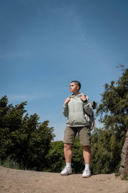 Young and confident short haired woman hiker in casual clothes holding backpack and looking away while standing with trees and blue sky at background, Translation of tattoo: love clipart