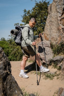 Young short haired female hiker with backpack holding trekking poles and looking at path while standing between stones with blue sky at background, trekking through rugged terrain, summer clipart