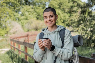Cheerful young short haired female hiker with fitness tracker and backpack holding thermos cup and looking at camera with landscape at background, finding serenity in nature, summer clipart