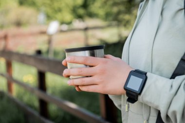 Cropped view of young woman tourist with fitness tracker holing cup of thermos and standing with blurred nature at background, finding serenity in nature, summer clipart