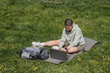 Young short haired and tattooed female tourist using laptop while sitting on fitness mat near backpack on grassy lawn with flowers, finding serenity in nature, summer clipart