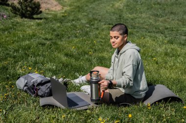 Genç, kısa saçlı, termos tutan bir kadın turist dizüstü bilgisayarın yanında fitness minderi ve çiçekli çimenlikte sırt çantasıyla otururken doğada, yazda, dijital göçebelerde huzur buluyor. 