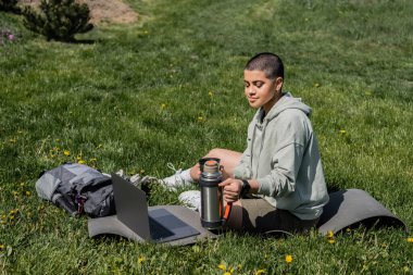 Young short haired woman tourist holding thermos while sitting on fitness mat near laptop and backpack on grassy lawn with flowers, finding serenity in nature, summer, digital nomad  clipart