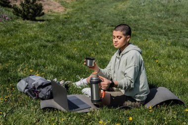 Young tattooed and short haired female hiker holding thermos while sitting near backpack and laptop on fitness mat on lawn with flowers, finding serenity in nature, summer, digital nomad  clipart