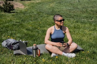 Young short haired female traveler in sunglasses holding thermos cup while sitting near laptop and backpack on fitness mat and lawn with flowers, finding serenity in nature, summer clipart