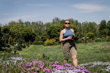 Young short haired woman tourist in sunglasses holding digital camera while standing near blurred flowers with blurred scenic landscape at background, Translation of tattoo: love clipart