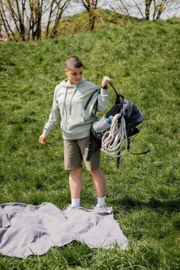 Young short haired female hiker with fitness tracker holding backpack with travel equipment while standing near blanket on grassy lawn with hill at background, connecting with nature concept clipart