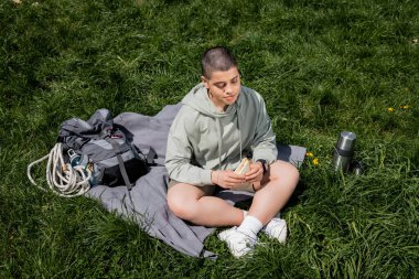 High angle view of young short haired female hiker holding sandwich while sitting near backpack and thermos on blanket and grassy lawn, connecting with nature concept, Translation of tattoo: love clipart
