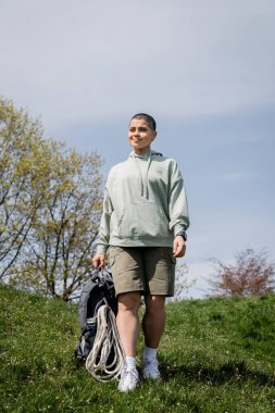 Gülümseyen, kısa saçlı, spor takip cihazıyla gezen, turizm ekipmanlarıyla sırt çantasıyla gezen, çimenli çimlerde yürüyen, manzaranın içinde yol alan, yaz mevsiminde