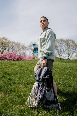 Kısa saçlı bayan gezgin, sırt çantasıyla seyahat ekipmanları ve spor şişesiyle arka plandaki çimenli çayırlara bakarken manzaranın içinde geziniyor.