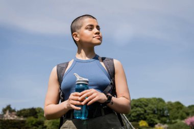 Young tattooed and short haired female hiker with backpack and smartwatch looking away and holding sports bottle with blue sky at background, trailblazing through scenic landscape, summer clipart