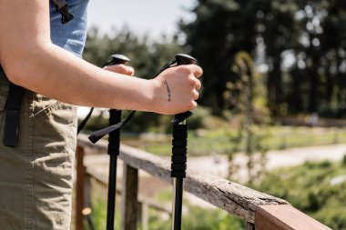 Cropped view of young tattooed woman tourist holding trekking poles while standing near wooden fence with nature at background, reconnecting with yourself in nature, Translation of tattoo: love clipart