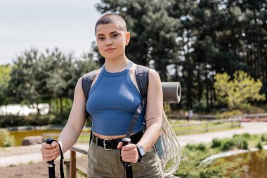 Portrait of young short haired and tattooed female tourist with backpack looking at camera while holding trekking poles with nature at background, reconnecting with yourself in nature concept clipart