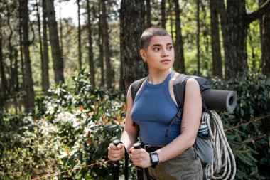 Young short haired female tourist with backpack holding trekking poles and looking away while standing in blurred green forest, reconnecting with yourself in nature concept, summer clipart