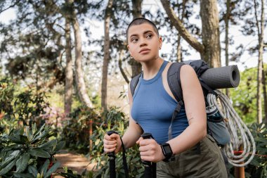Young short haired and tattooed female hiker with backpack holding trekking poles while looking away and standing in blurred summer forest, hiking for health and wellness concept  clipart