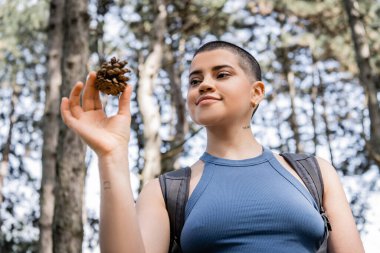 Low angle view of young tattooed and short haired female tourist with backpack holding pine cone while standing in blurred summer forest, hiking for health and wellness concept  clipart