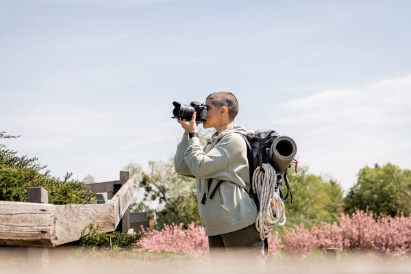 Вид сбоку на молодого коротковолосого и татуированного туриста с рюкзаком, фотографирующего на цифровую камеру, стоя возле деревянного забора и живописного ландшафта, путешествующего фотографа