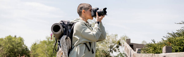Вид сбоку на молодую татуированную и коротковолосую туристку с рюкзаком, фотографирующуюся на цифровую камеру, стоя возле деревянного забора на природе, пешеходный поход по ландшафту, баннер 