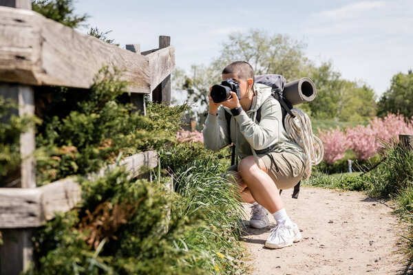 Молодая коротковолосая туристка с рюкзаком и туристическим оборудованием, фотографирующая на цифровую камеру возле деревянного забора и траву на заднем плане, путешествующий фотограф 