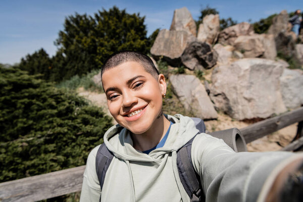 Portrait of cheerful tattooed and short haired female tourist with backpack looking at camera while standing with blurred scenic landscape at background, exploring new horizons, summer