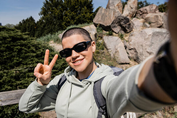 Smiling short haired and tattooed female tourist in sunglasses with backpack showing peace gesture while standing with scenic landscape at background, exploring new horizons, summer