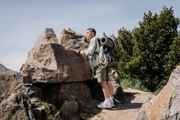 年轻的短发女徒步旅行者 背着背包和旅行装备 站在石头旁边 与自然和天空相映成趣 探索新的地平线 — 图库照片