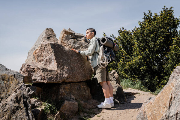 Young short haired female hiker with backpack and travel equipment looking away while standing near stones on path with nature and sky at background, exploring new horizons, summer