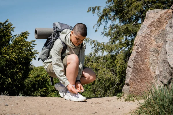 stock image Young short haired female traveler with backpack and fitness mat tying shoelace on sneaker and standing near stones on path with nature at background, trekking through rugged terrain