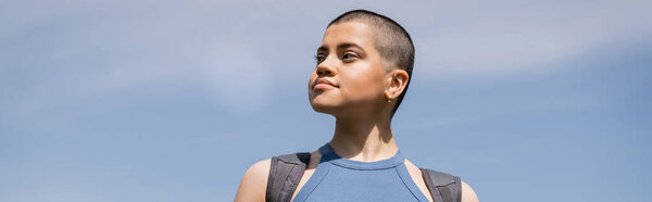 Young confident and short haired female tourist with backpack looking away while standing with blue sky at background, solo hiking journey concept, banner, summer