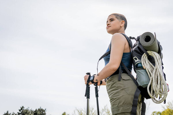 Low angle view of young short haired female backpacker with fitness tracker and backpack looking away while holding trekking poles with sky at background, solo hiking journey concept 