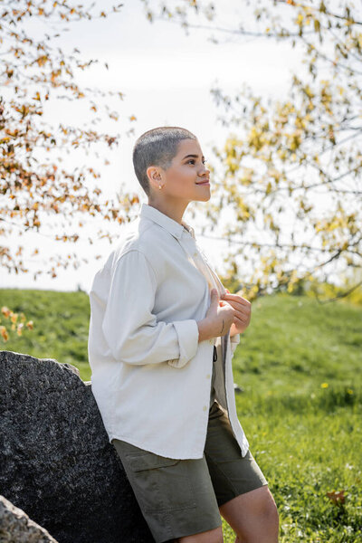 Positive young short haired female hiker wearing casual shirt while looking away and standing near stones with blurred scenic landscape at background, Translation of tattoo: love