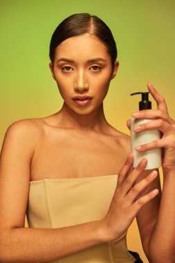 product presentation, skin care product, young asian woman with bare shoulders holding cosmetic bottle with body lotion and posing on green background, glowing skin concept  clipart