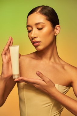 Cilt bakımı, ürün sunumu, genç cilt, çıplak omuzlu genç Asyalı kadın kozmetik tüpü tutuyor ve yeşil arka planda poz veriyor, parlayan cilt, esmer saç 