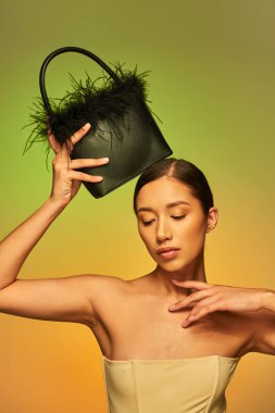 beauty and style, brunette asian woman with bare shoulders posing with feather purse on green background, hand near face, gradient, fashion statement, glowing skin, natural beauty, young model  clipart