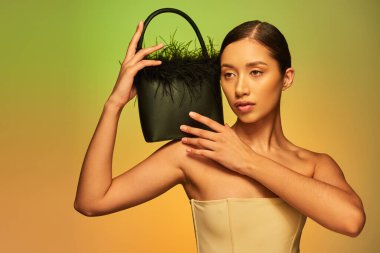 Moda seçimleri, çıplak omuzlu esmer Asyalı kadın. Tüylü çanta ve yeşil arka plana bakıyor. Gradyan, moda anlayışı, parıldayan ten, doğal güzellik, genç model. 