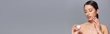 Cilt bakımı, esmer saçlı genç Asyalı kadın kozmetik kavanozu tutuyor ve gri arka plana yüz kremi sürüyor. Parlak ve sağlıklı cilt, güzellik kampanyası, yüz bakımı konsepti, afiş. 