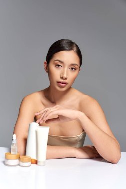 beauty photography, young asian woman with brunette hair posing near beauty products on grey background, glowing and heathy skin, facial treatment concept, facial and skin care, youth  clipart