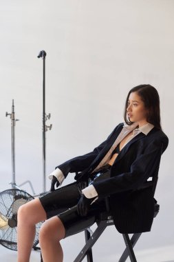 studio photography, young asian model in blazer, white shirt and latex shorts sitting on folding chair near electric fan on grey background, fashion and style, looking away, full length  clipart