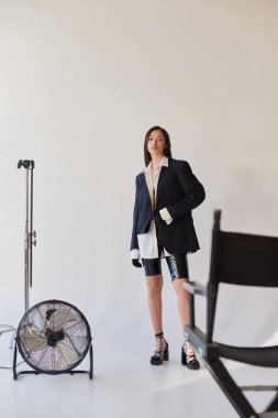 Cesur stil, stüdyo fotoğrafçılığı, şık görünümlü genç Asyalı kadın gri arka planda poz veriyor, ceket, beyaz gömlek ve lateks şort, elektrikli yelpazenin yanında duruyor, kişisel moda seçimi, tam boy. 