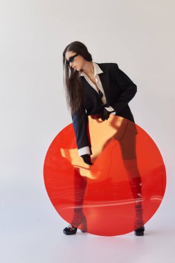 Şık görünümlü ve güneş gözlüklü çekici Asyalı model kırmızı yuvarlak cam, gri arka plan, ceket ve lateks şort, genç ve modern kadın, kenar stili, stüdyo fotoğrafçılığı 