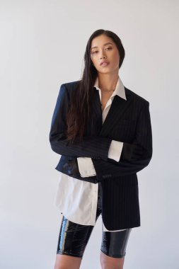 Moda ileri, modaya uygun genç Asyalı kadın lateks şortlu, gri arka planda poz veren, ceket ve siyah eldivenli, katlanmış kollarıyla duran, genç model, stüdyo fotoğrafçısı, kavramsal 